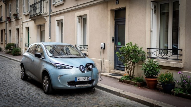 Voiture Ă©lectrique compacte dâoccasion en recharge devant une maison française, entourĂ©e de vĂ©gĂ©tation urbaine dans une ambiance rĂ©aliste et sobre.